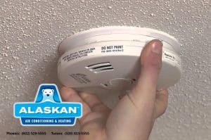 hand holding fire detector near ceiling Alaskan Air Conditioning and Heating logo and contact phone numbers