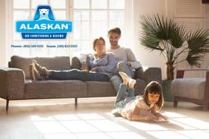 Couple sitting on couch while child lays on floor interacting with tablet Alaskan Air Conditioning and Heating logo and contact phone numbers in top left quadrant