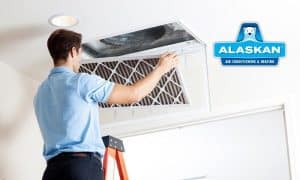 man standing on ladder to remove air filter from ceiling vent with Alaskan Air Conditioning and Heating logo