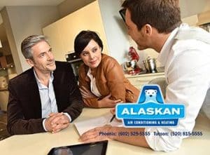 couple sitting at table looking at man in white shirt (3/4 rear view from camera) who holds pen and paper, Alaskan Air Conditioning and Heating logo and contact phone numbers in bottom right corner